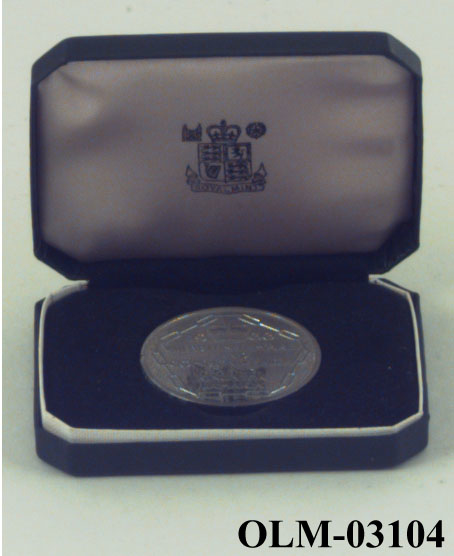 Sølvmynt med en kjede langs kanten, en krone øverst og tekst i midten. På baksiden en kvinne med fruktkurv og en mann med bue, mellom dem et våpenskjold med en krokodille øverst. Mynten ligger i en eske.