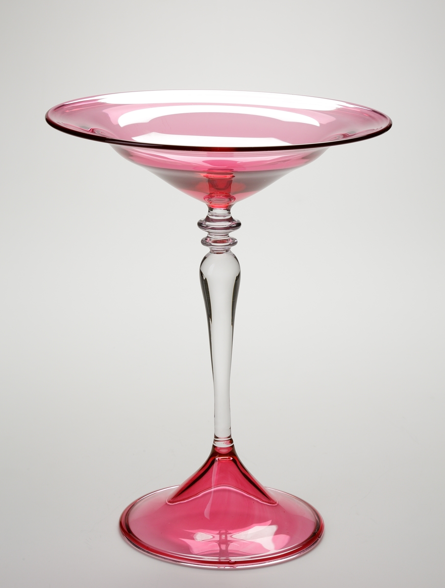 Skål på ben och fot.
Stilen på glaset är gjord så att den ska se ålderdomlig ut.
Skålen i rosa är kraftigt utvikt. 
Det ofärgade balusterformade benet har överst två knappar.  
Foten rosa.
