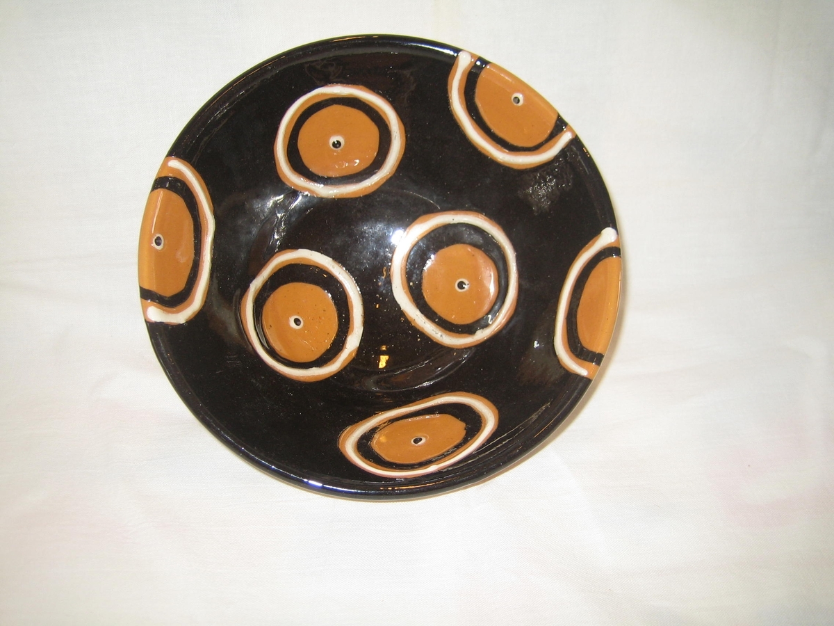 Rund skål i naturfärgad keramik på utsidan och svart glasyr på insidan, dekorerad med cirklar i vitt och svart med en vitsvart prick i mitten.