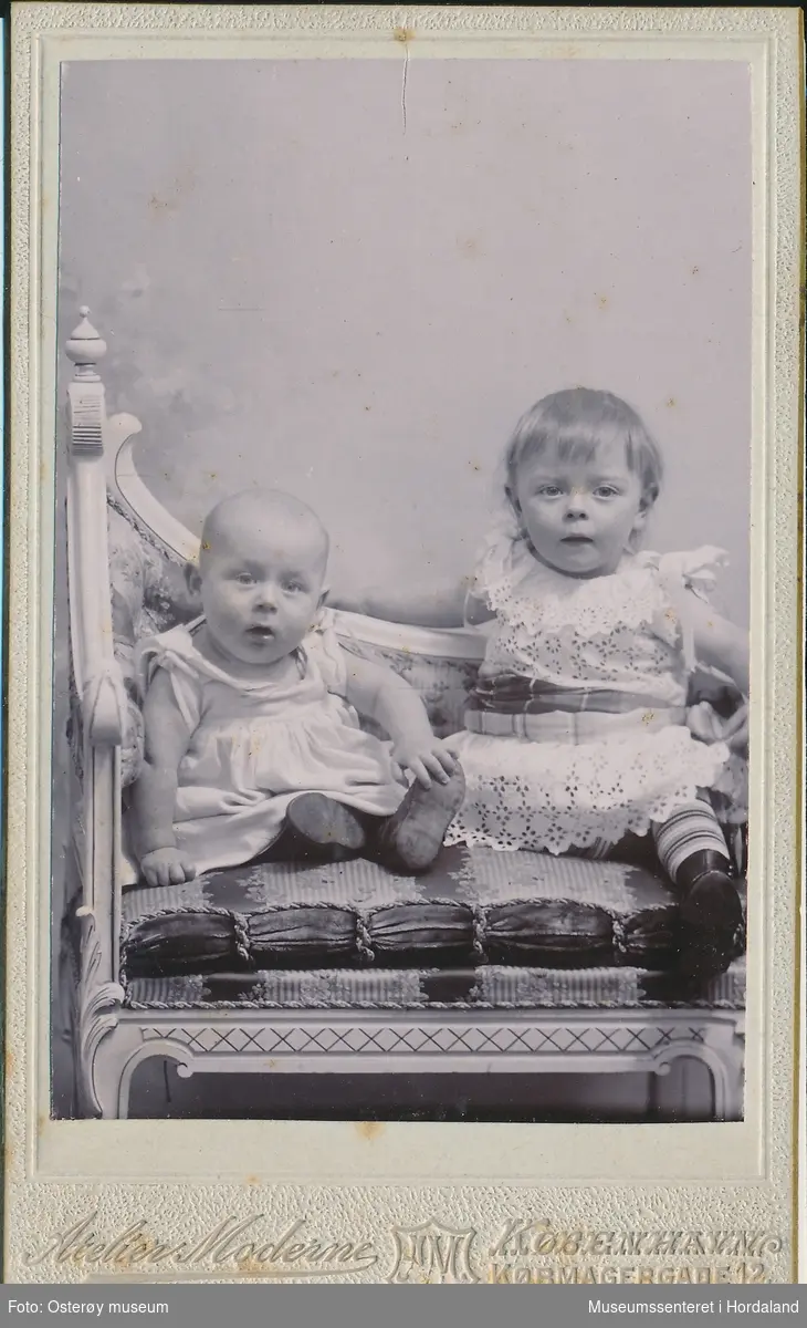 atelierfotografi av to små born, gut og jente som sit i ein stol kledd i ljose kjoler