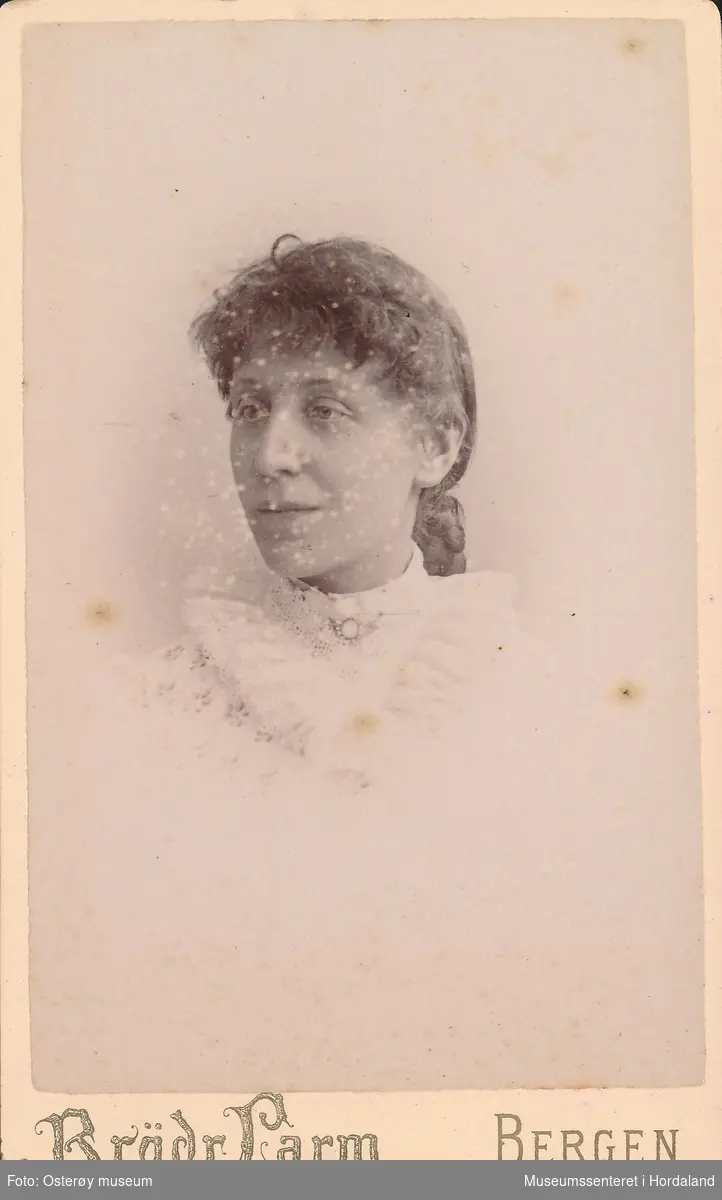 portrettfotografi av kvinne med pannelugg, hårknute i nakken, kvit blondebluse med stor krage i engelsk broderi og nål i halsen