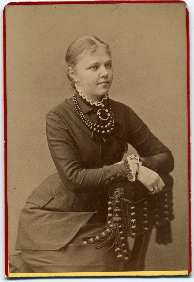 Kabinettsfotografi: okänd ung kvinna med halsband och brosch, sittande med armarna vilande på stolsryggen.
