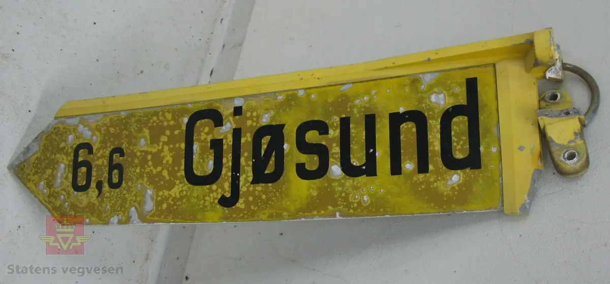 Vegviserskilt. Skiltplata er lysreflekterende gul med svart tekst "Gjøsund 6,6". Selve skiltplata er lagd av en aluminiumsplate der festet og kanten øverst er støpt i en del. Skiltet har vært festet til 3 toms stolpe ved hjelp av to bøyler. En bøyle henger på skiltet
