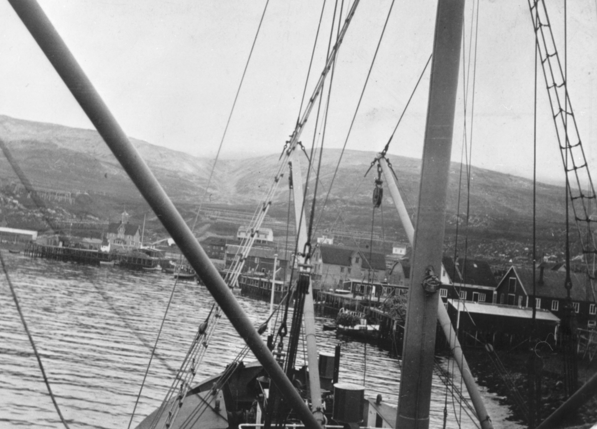 Bildet viser Båtsfjord havneområde med båter, kaier, pakkhus og rorbuer, i bakgrunnen fiskehjeller. Bildet er fotografert fra en båt.