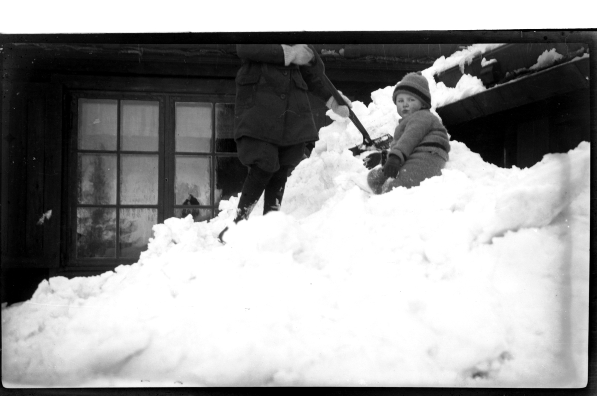 Julius Sundt sitter i snøen mens en mann måker snø, Villa Knyggen. Fotografert 1919.