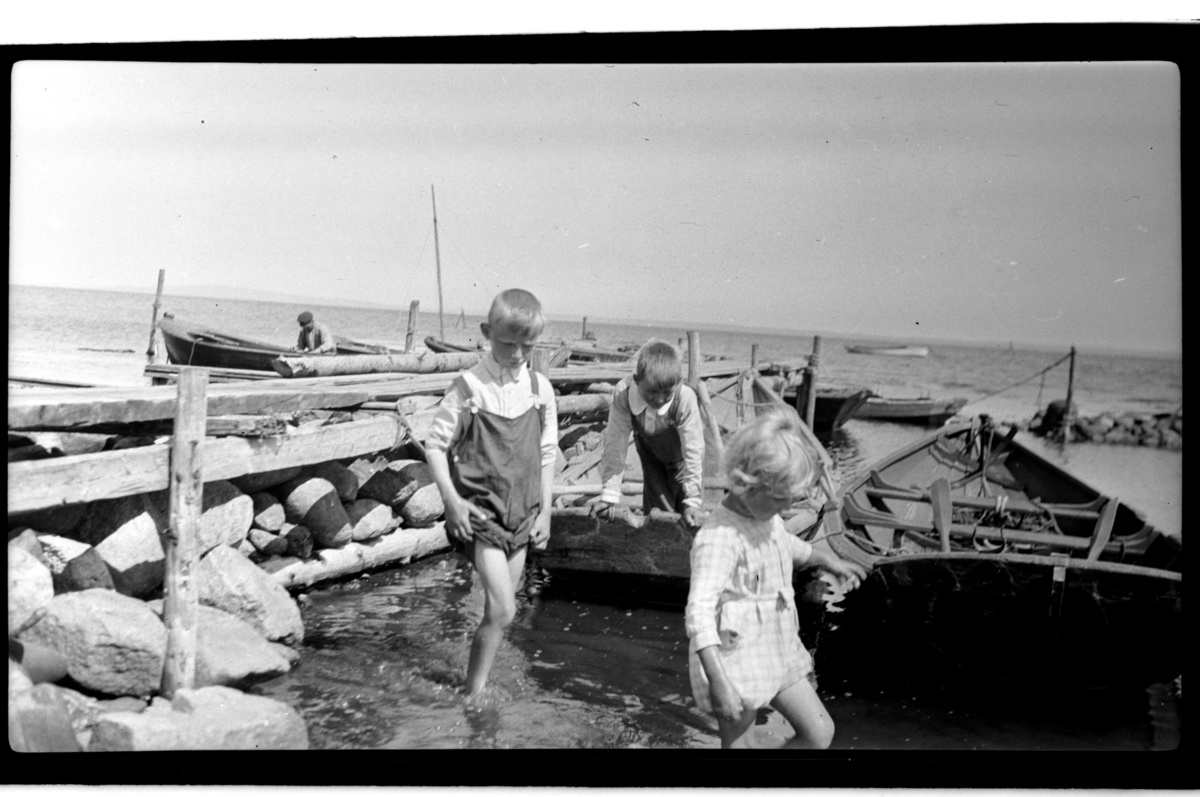 Julius Sundt, en ukjent gutt og Rolf Sundt jr. vasser ved en brygge, flere robåter ligger fortøyd, Slagen. Fotografert 1925.