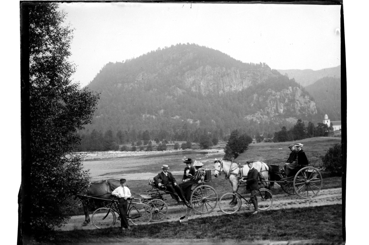 To hestekjøretøy med passasjerer og to syklister i ukjent landskap med kirke. Fotografert rundt 1902.