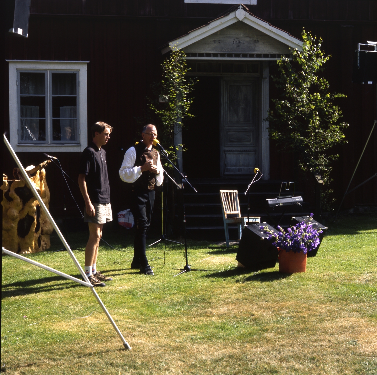 Rengsjöfesten 11 juli 1999. En man klädd i hembygdsdräkt talar i en mikrofon och bredvid honom står en yngre man. Det delas ut belöningar. Där finns en talarstol och en högtalaranläggning.