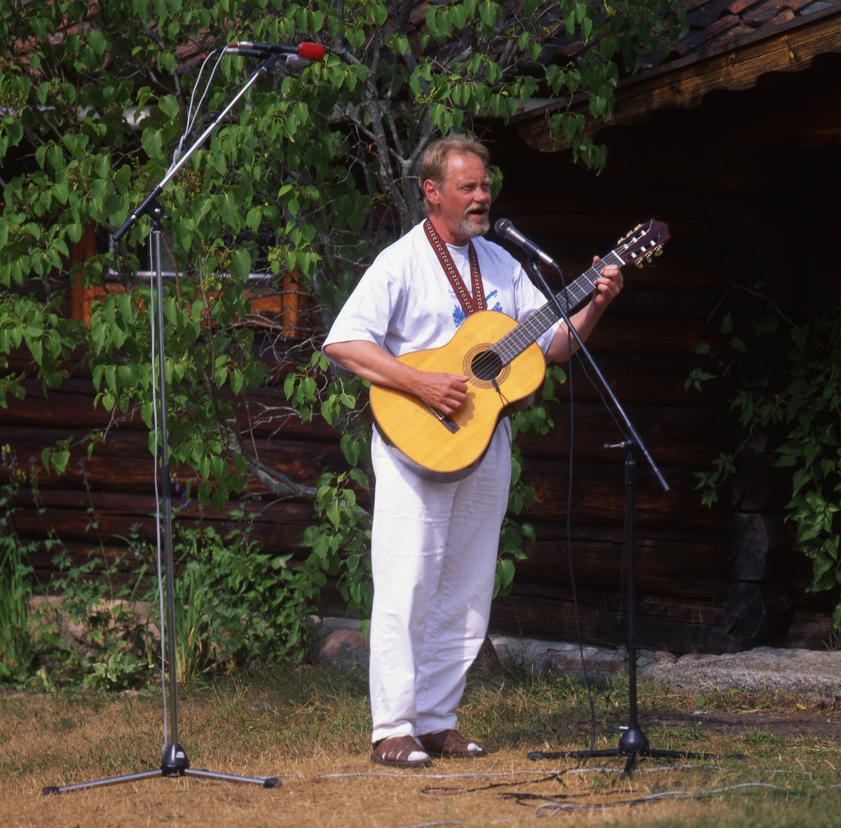 En man står utomhus framför en timrad byggnad och spelar gitarr och sjunger. Två mikrofoner är uppställda intill honom.