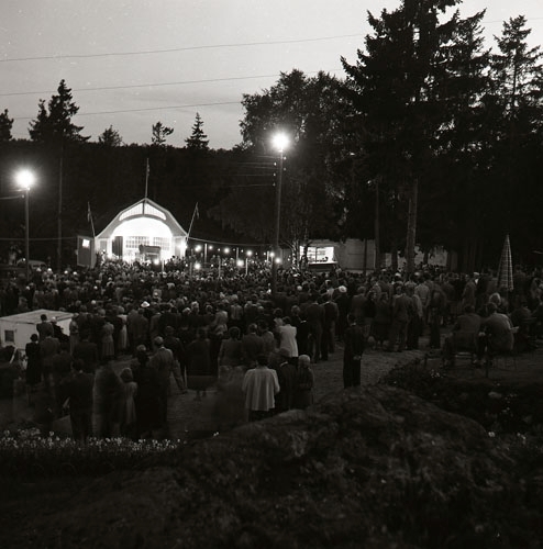 Kvällsfest på "berget" i Söderhamn augusti 1953. En man står framför en mikrofon på en upplyst utescen med skylten SGU. Framför sitter en stor publik.