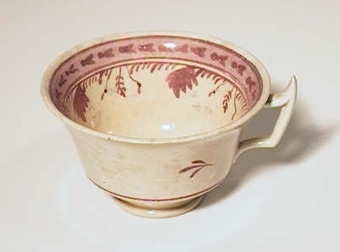 Vit kopp av flintgods. Dekor med bårder, blad och blomslingor i rosa. Lysterglaserad.
