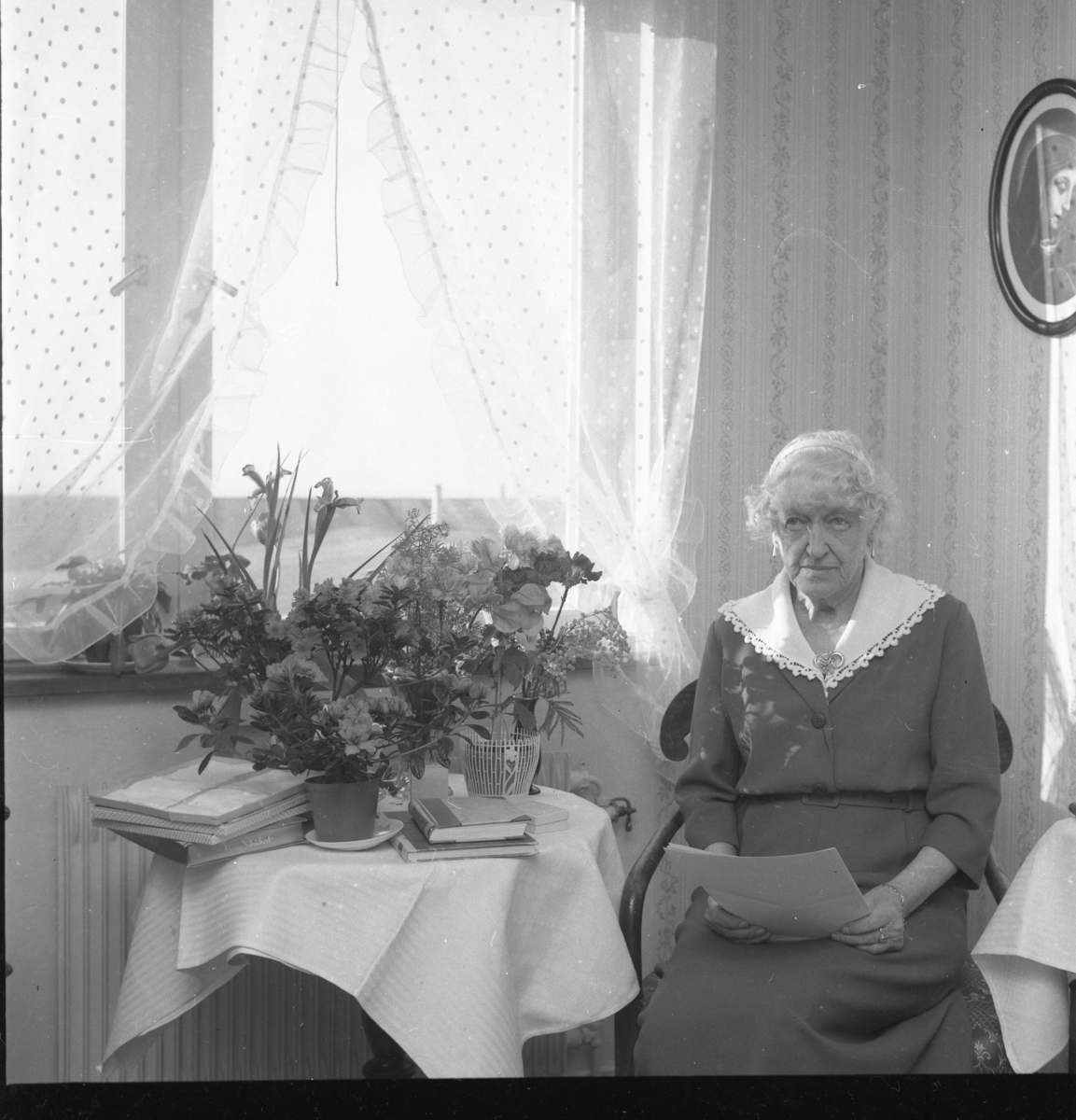 Fröken Hård af Segerstad sitter vid ett bord med blommor och håller några papper i knät. Hon heter eventuellt Gertrud och fyller 80 år.