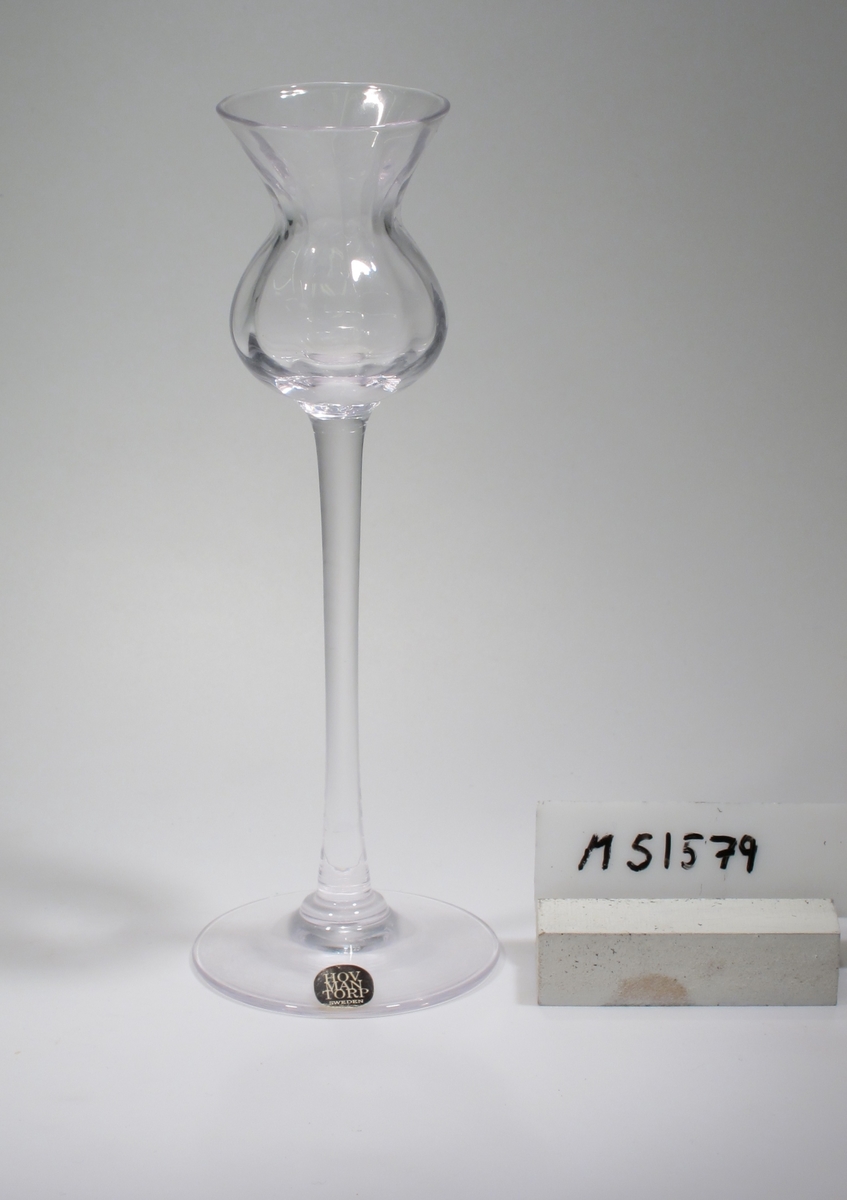 Ljusstake, klockformad kupa med stolpoptik, högt ben.
Etikett: Svart/vit: ”HOVMANTORP SWEDEN”