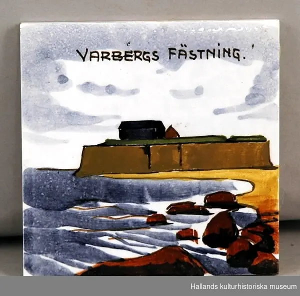 Kakelplatta med bild av Varbergs fästning och texten: "VARBERGS FÄSTNING HANDMÅLAT". Stämpel på baksidan: "MADE IN BELGIUM".