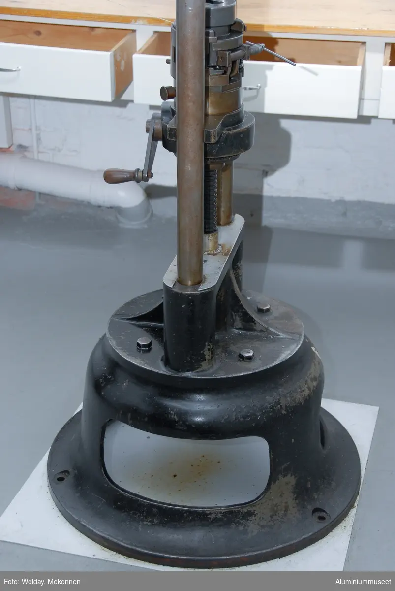 Strekkprøvemaskin for metaller.  Leverandør: Alfred J. Amsler & Co. Schweiz.  Datering: ca. 1925-1966. Teknikk: Hovedenhetene er støpt i stål og delvis maskinert.  Enkelte deler er fremstilt av valset stål og hydrauliske overføringer er kobber-rør.
Form: To stk. maskinenheter.  En betjeningsenhet og en strekkenhet som er forbundet med hydrauliske rør.
Tilstand: Maskinen er operativ, men har hydrauliske lekasjer.
Utfyllende opplysninger:
Betjeningsenheten består av en hydraulisk pumpe som via 2 stk. reguleringsventiler leverer olje til sylinderen for pendelmanometeret og til hovedsylinderen på toppen av strekkenheten.  Strekkprøvene plasseres i klemmebakkene på strekkenheten, blyanten settes på papirrullen og høyre kran på betjeningsenheten åpnes.  Etterhvert som trykket økes blir prøven strukket helt til brudd.  Under strekket slår pendelen ut tilsvarende trykket i hovedsylinderen og strekk-kraften kan til enhver tid avleses på skala-skiven.  Via et snorsystem fra overbjelken hvor øvre klemmbakke sitter, vil strekkforløpet bli avtegnet på en papir-rull på betjeningsenheten og strekkforløpet blir avtegnet slik at hele strekkforløpet og flytgrensen kan avleses.  Ved bruk avstenges hovedkranen umiddelbart og da vil den sorte pila umiddelbart gå tilbake til null-posisjon., men den røde pila blir stående igjen og viser bruddkraften.  Med den venstre kranen slippes hovedsylinderen ned til utgangsposisjon igjen.  Tykkelse og bredde på prøvestaven blir målt før prøvetaking og staven blir også risset med tynne paralelle strekker med 10 mm mellomrom.  Se risseapparat HAM.79.  Etter brudd blir prøvestaven satt sammen igjen og målt over en lengde på 5 riss med bruddet ca. midt i mellom. Den nye lengden i forhold til 50 mm viser forlengensen som blir utregnet i prosent.  Bruddfastheten er avlest bruddkraft delt på tverrsnitt.  Flytegrensen avleses av det tegende diagrammet på papir-rullen pluss 0,2%.  Maskinen kan maks. yte 10 tonn, men pga. unøyaktig avlesing ved tynne prøver blir pendelmanometeret innstilt ettter forventet bruddstyrke.  Maskinen har 4 innstillnger: Maks.: 1000 kg: Nest nederste hull. Uten lodd. Maks.: 2000 kg: Nest øverste hull. Uten lodd. Maks.: 5000 kg: Nederste hull.  Med Lodd. Maks.: 10000 kg.: Nest øverste hull. Med lodd.
Maskinen står i dag 08.01.2007 i psosisjon, 5000 kg.  Maskinen kan også brukes til trykkprøving, f.eks. sveiseprøver, ved å plassere prøven på rullebakkene oppe på strekk-bommen.  22.01.2008 Kjell Grette.