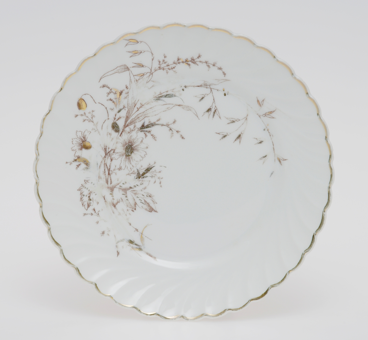 Sirkulær tallerken i porselen med glasur. Dekorert med en gullforgylt ring rundt kanten som er bølgete. Blomster- og bladmotiv på halve tallerkenen. Skråstilte riller støpt inn i porselenet.