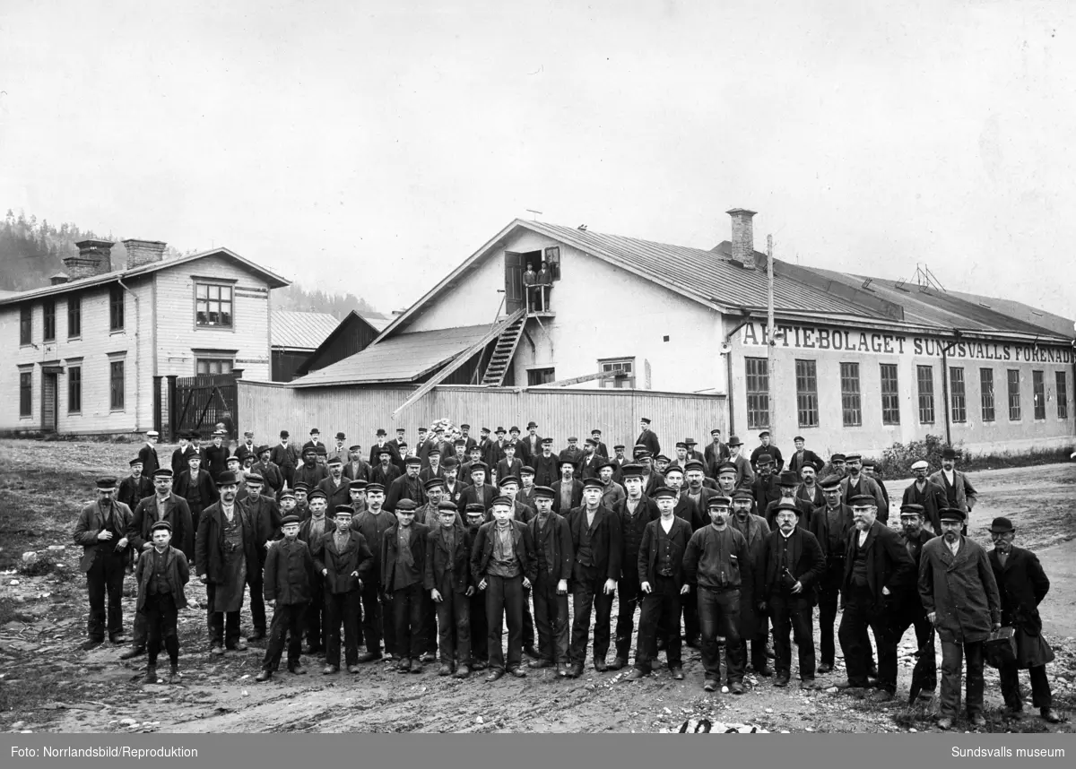Reproduktion av ett gruppfoto från 1906 med arbetare vid Aktiebolaget Sundsvalls Förenade Verkstäder. Bilden är tagen vid dåvarande byggnadens västra ände.