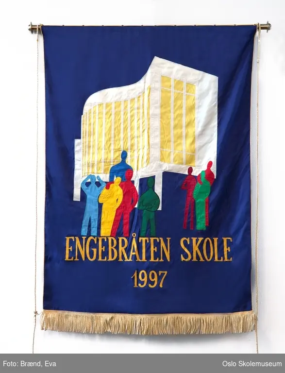 En gruppe menneske silhouetter i primær farger foran en stilisert bygning (skolen) på blå bakgrunn. Nederst på fanen står skolens navn og grunnleggelses år i gull.
