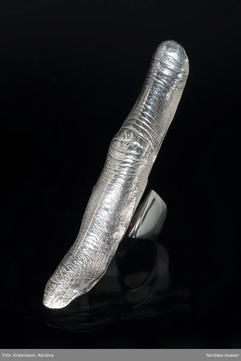 Ring av silver, överdel i form av avlång avgjutning av fingerrygg. Underdel bestående bred ring med slits på ena sidan. Undertill graverad "Cornelia".
/Leif Wallin 2018-02-09