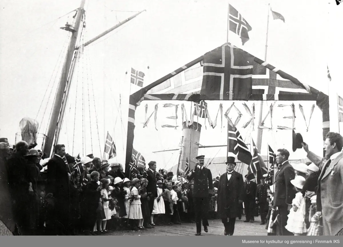 Kong Haakon VII ankommer Honningsvåg. Han passerer sammen med John Bruun velkomstportalen og ble møtt med flagg og hurra-rop. 28.06.1922.