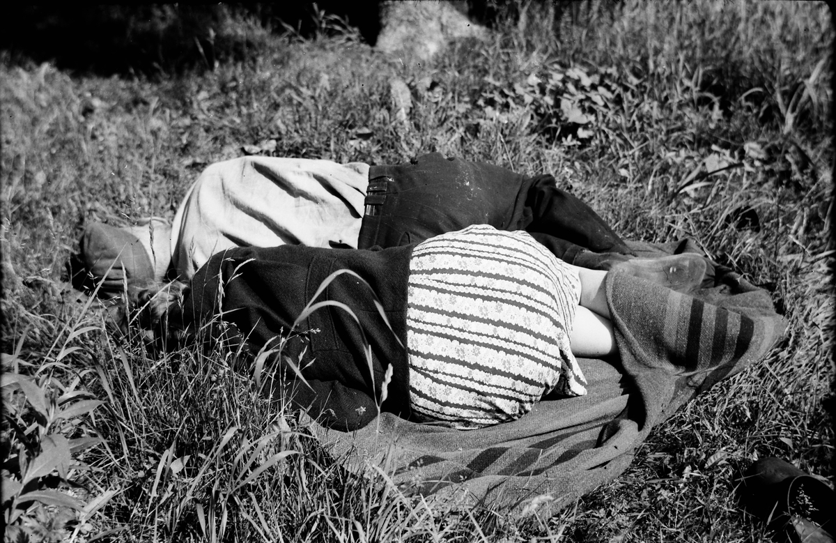 Ett par vilar i gräset på en filt. Det tycks vara lite kyligt för kvinnan har vikt upp filten över vaderna.
