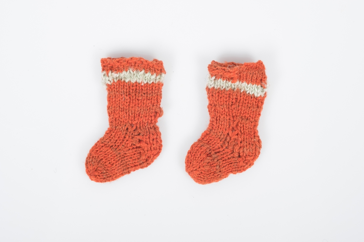 Et par bittesmå strikkede ullsokker. Sokkene er oransjerøde med en lys stripe øverst og nøen brunrøde striper lenger ned.