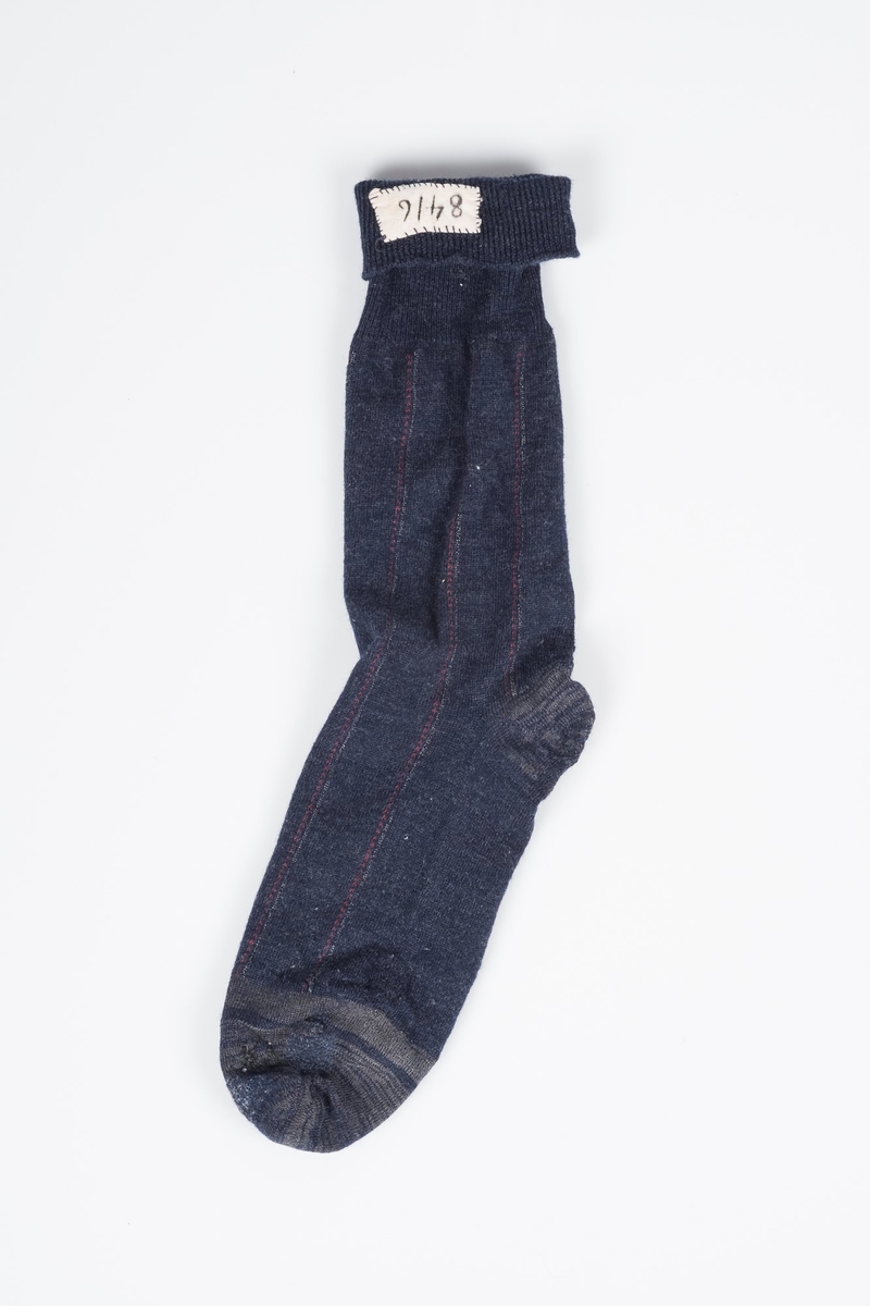Mørkeblå høy sokk, med grå detaljer og røde og hvite striper. Sokken er meget godt brukt og stoppet flere ganger. På innsiden øverst er det sydd inn en lapp med fangenummeret 8416, som identifiserer sokken som Erich Mønnichen Plahtes.