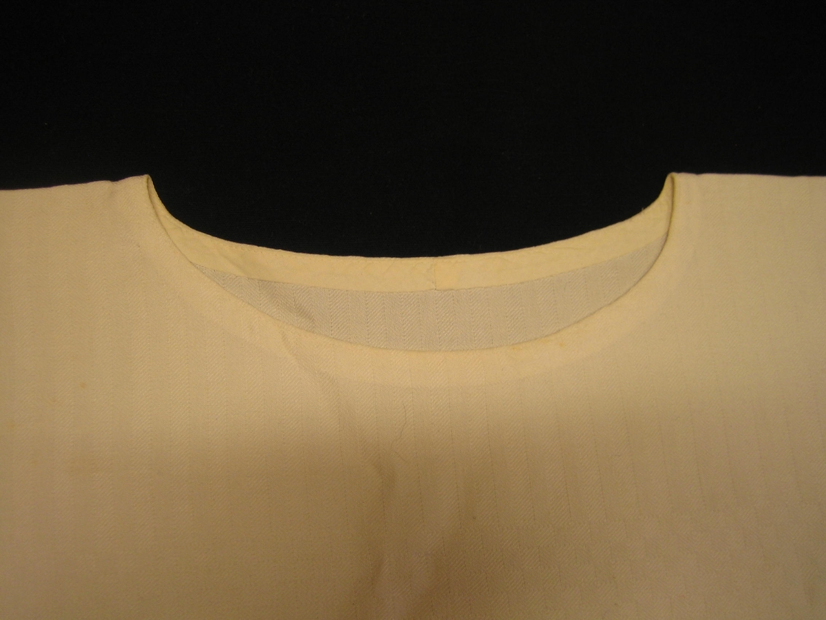 Vit blus i halvlinne med mönster i rutig och randig kypert. Blusen är försedd med oval halslinning som är nedsydd med en snedremsa. Den är försedd med 10 cm långa sprund i sidorna. Ärmarna är rakt isydda vid fram- och bakstycke och försedda med en kil i ärmhålan. Tyget är beskrivet i RSLH-0081.