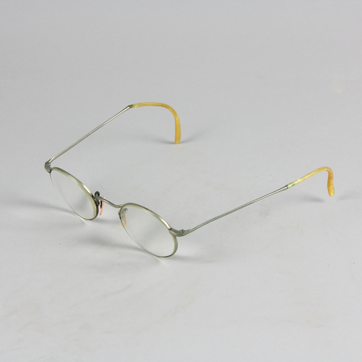 Glasögon med runda glas infattade i metall och böjda skalmar av metall som avslutas med annat material i gulvitt. Näskuddar i gulvitt respektive rosa.