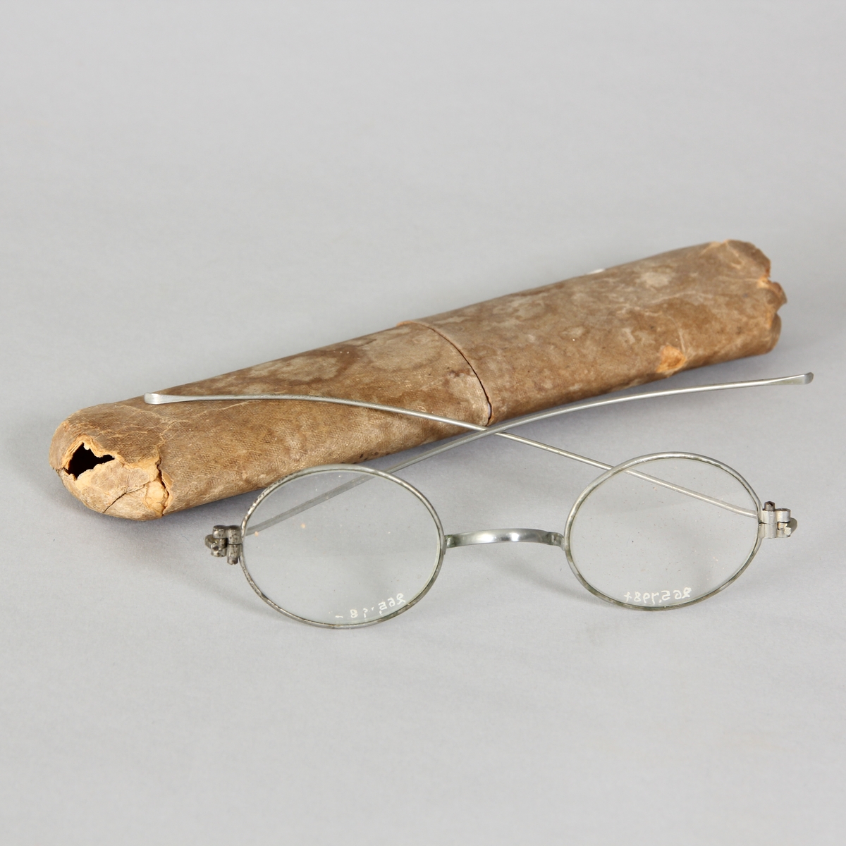Glasögon, 1 par, med ovala glas och tunna vitmetallskalmar. Avlångt fodral av papp med rundade hörn, delbart.