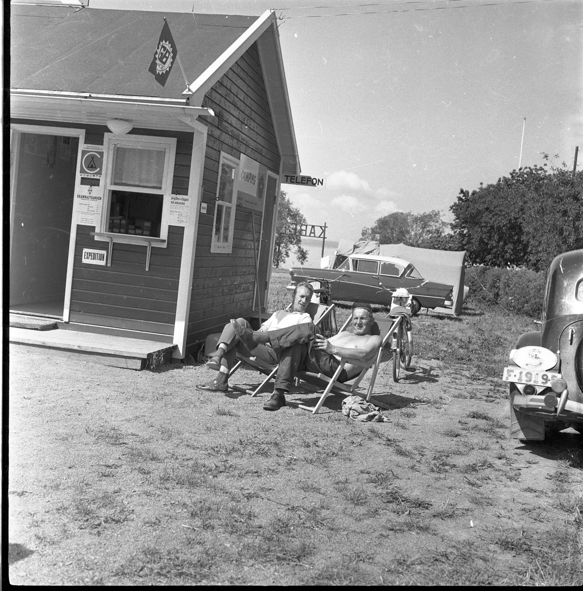 Ernst Callas och Ivar Fager (med bar överkropp) sitter i varsin solstol intill Gränna campings expedition. Bakom dem står en Opel intill ett tält och till höger skymtar en Citroën.