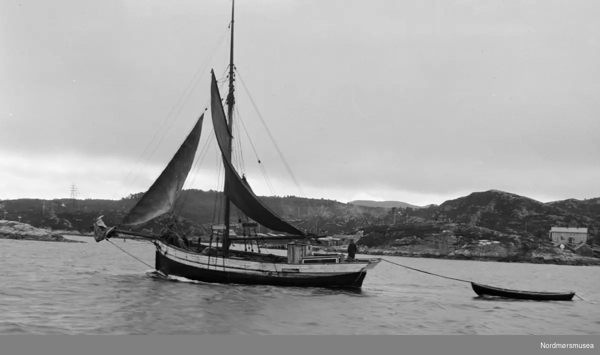 Foto av en seilbåt/fiskeskøyte med en liten robåt på slep. Trolig fra Eidsvåg i Nesset kommune, med Georg Sverdrup som sannsynlig fotograf. Fra Sverdrupsamlingen ved Nordmøre museums fotosamlinger.
