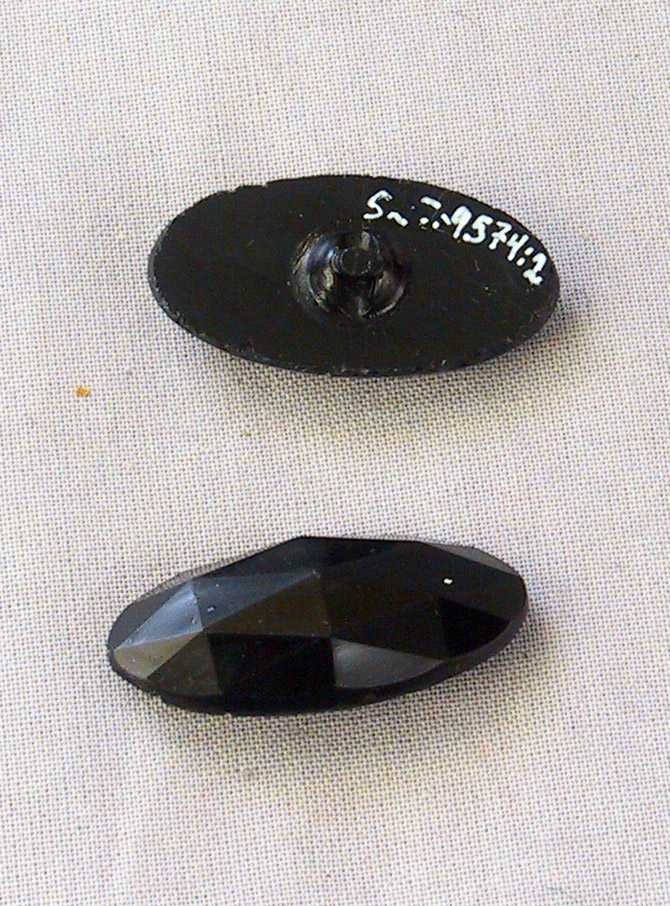 Oval, svart plastknapp med trekanta fasettar oppå. Sirkulær tapp med hol gjennom på undersida.
