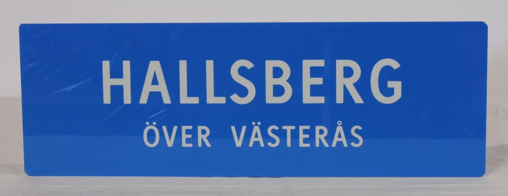 Destinationsskylt av ljusblå plast med texten "HALLSBERG ÖVER VÄSTERÅS" i vitt. Spegelvänd text på baksidan.
