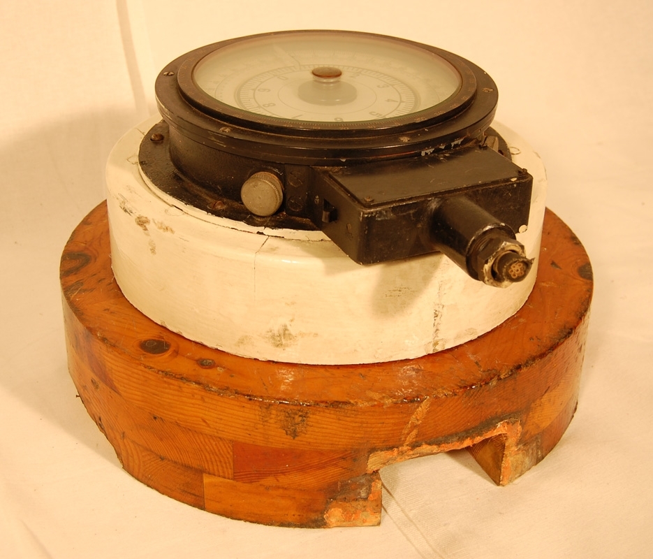 Gyrokompass som använts på en av SJ:s färjor, okänt vilken. Kompassen är fastskruvad på en vitmålad träcylinder. Den vitmålade träcylindern har i sin tur varit fästad i en lackad träcylinder, nu ligger den löst ovanpå.