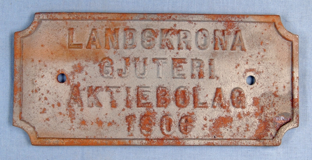Rektangulär liggande skylt med avkapade hörn, brun. Text i relief: "LANDSKRONA GJUTERI AKTIEBOLAG 1906".