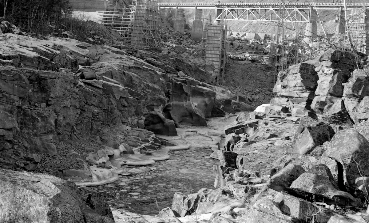 Anleggsarbeid i forbindelse med bygginga av et kraftverk ved Harpefossen i Gudbrandsdalslågen, i Sør-Fron kommune i Oppland fylke, i 1964.  Fotografiet er tatt mot et nesten tørrlagt elveleie i et trangt parti med bratte, opprevne bergskrenter på begge sider av elveløpet.  Da dette fotografiet ble tatt var man i ferd med å forskale og støpe betongfundamenter, som antakelig skulle inngå i en damkonstruksjon.  I bakgrunnen (øverst i bildeflata) ser vi ei stålbru på betongfundamenter, med fagverksunderstøtting under det langt midtspennet.  Under dette spennet skimter vi en skådam av armert betong som ble støpt for å lede hovedstrømmen i elva mot inntaktspunktet for kraftverkets turbiner.  Kraftstasjonen ved Harpefossen har to kaplanturbiner og en årsproduksjon på 427 GWh.  Anlegget utnytter et fall på 34 meter i Gudbrandsdalslågen.  Det ble satt i drift i 1965.