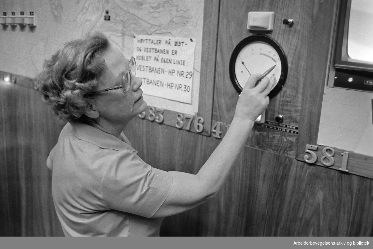 Drosjesentralen. Vaktsjef Ragnhild Bekkby på drosjesentralen viser fram meteren som viser pågangen av telefoner. Desember 1975.
