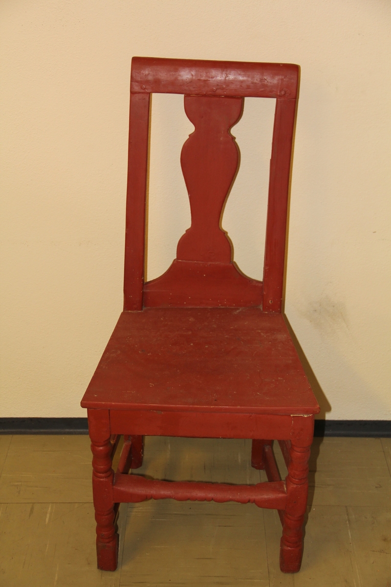 Rødbrun stolpekontruert trestol med dreide bein. Stolen er dekorert med utskjæring i ryggbrettet og midtsprosse