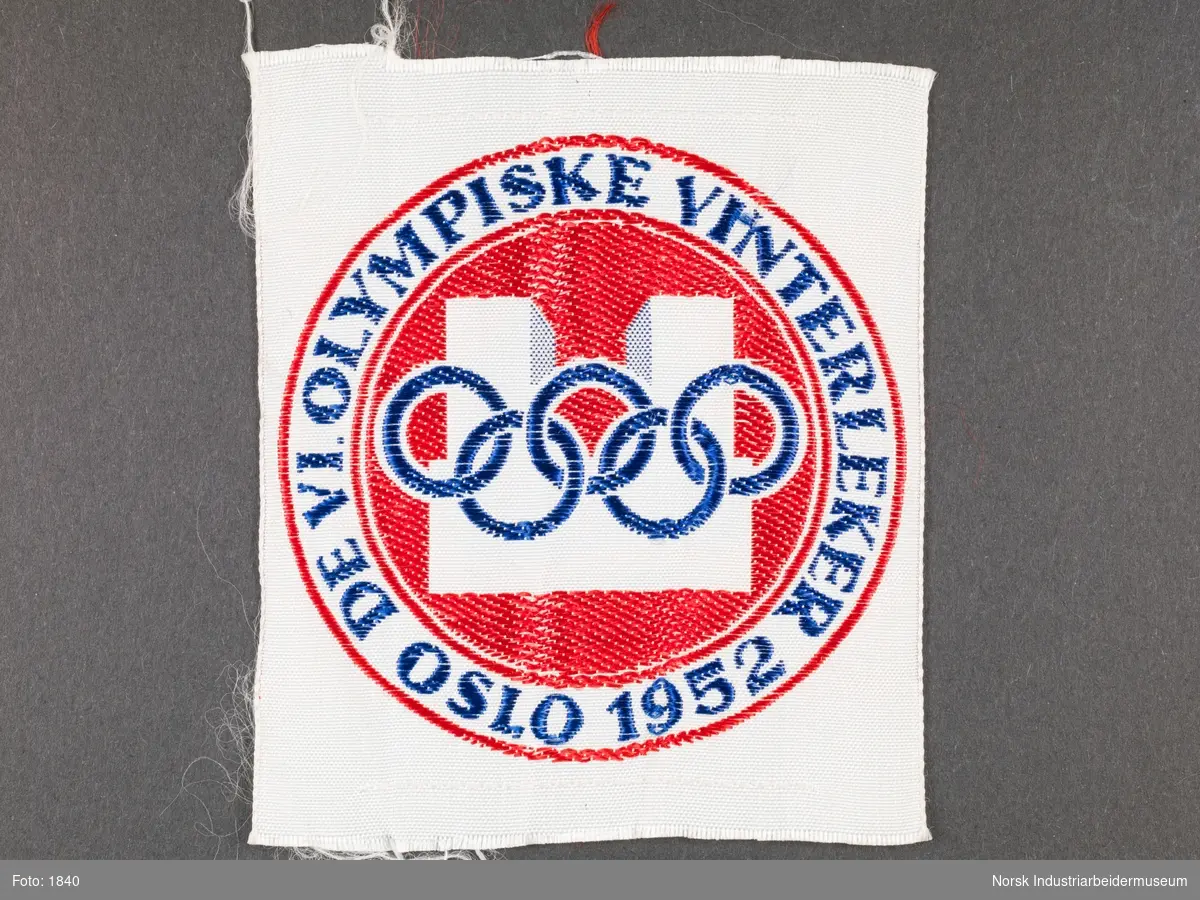 Kvadratformet tøystykke/merke. Sirkelformet logo med Oslo Rådhus og de olympiske ringer i midten. I sirkel rundt står det "De VI Olympiske Vinterleker Oslo 1952".