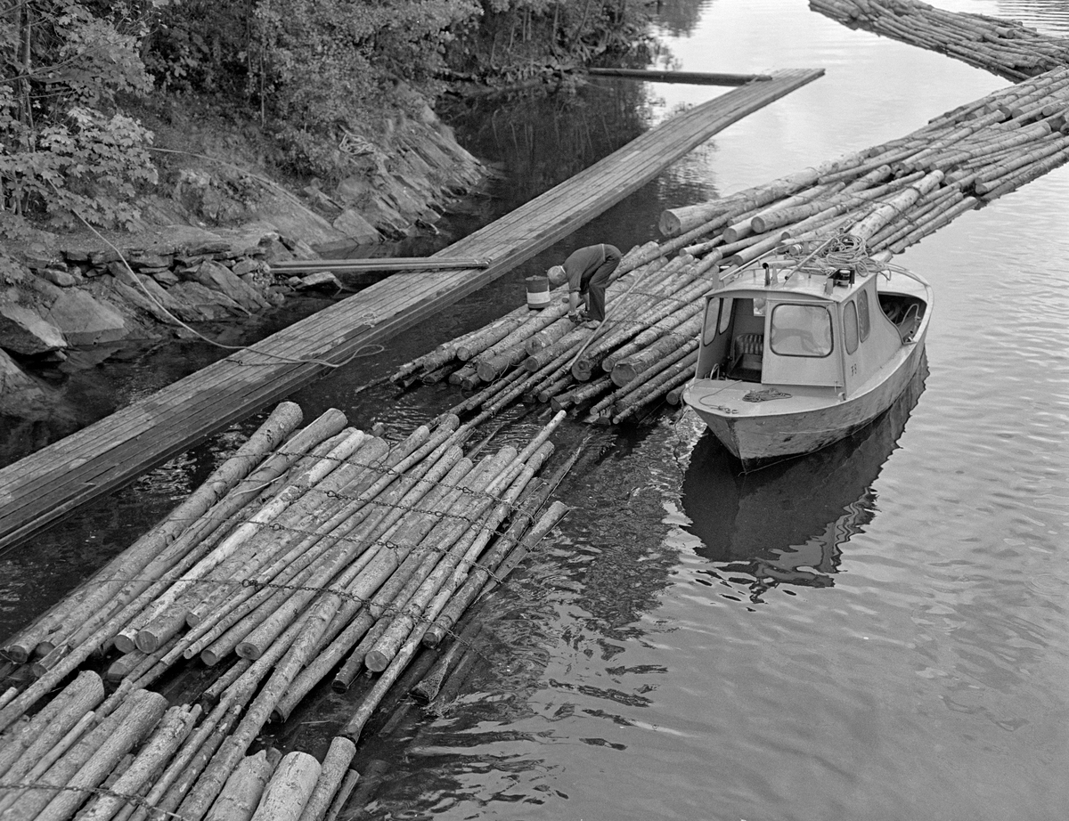 Fra kanalen nedenfor Strømsfoss sluse i Aremark i Østfold.  Fotografiet er tatt i 1982, som var den siste sesongen med tømmerfløting i Haldenvassdraget.  De siste tre åra ble det utelukkende levert ubarket massevirke i tre meters lengder.  Dette tømmeret ble utislått som bunter fra lastebil høyere oppe i vassdraget.  Buntene ble samlet i store slep, som ble trukket over innsjøene ved hjelp av kraftige slepebåter.  På slusestedene måtte imidlertid slepene deles opp i kortere lenker som passet inn i slusekamrene.  Av det nevnte sortimentet gikk det fire bunter i hver slusevending.  Nedenfor slusene, som her på Strømsfoss, ble disse slusevendingene bundet sammen i lengre lenker igjen.  Og lenkene ble i sin tur bundet til større slep på et flåtested i nordenden av Aremarksjøen.  Her ser vi pensjonert slusemester Odd Johansen i arbeid på ei tømmerlenke som lå inntil gangbanen på østre side av kanalen nedenfor Strømsfoss lense.  Sannsynligvis skulle han kople endeflatene på to slusevendinger til hverandre ved å trekke et par stokker fra endebunten på den ene over på endebunten på den andre og smøye disse innunder vaierbindene, der de ble festet med ståltrådkramper over vaierne. Ved siden av tømmerbunten Johansen arbeidet på ser vi en av de 18 varpebåtene Haldenvassdragets Fellesfløtningsforening disponerte.  Det var en stålbåt der den ene halvdelen av styrhusfronten kunne slås opp som ei dør, slik at mannskapet lett kunne ta seg fram på det lille fordekket uten fare for å falle i vannet. 

En liten historikk om tømmerfløting og kanaliseringsarbeid i Haldenvassdraget finnes under fanen «Opplysninger».