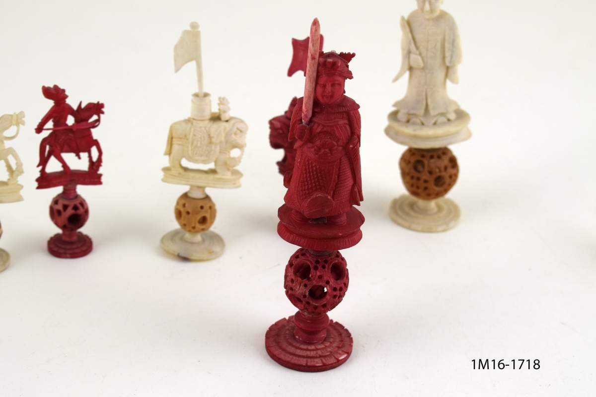 Röda och vita shackpjäser. Totalt 32 figurer. Figurerna i form av människor och elefanter. Minsta figuren 6cm. Största figuren 13 cm.