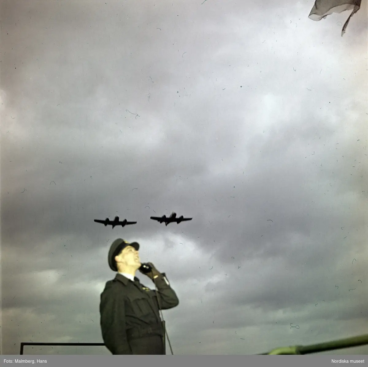 Brittiska flygvapnet, Royal Air Force (RAF). En flygvapenofficer talar i telefon under mörka skyar. Ovanför honom flyger jaktplan av typen Gloster Meteor F.4. Troligen flygbasen Duxford i Cambridgeshire, England.