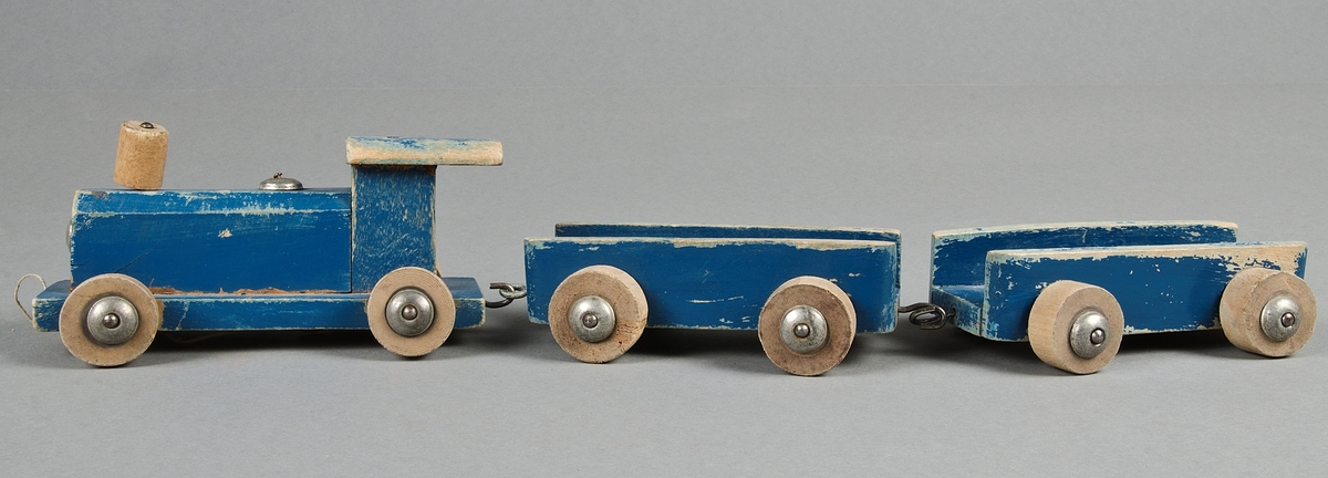 Leksakståg av trä, blåmålat med omålade trähjul. ett lok och två öppna vagnar som kopplas ihop. Metallkrokar och axlar och brickor på hjulen. Vitt snöre att dra det i. Färgen nött och hjulen vinda.