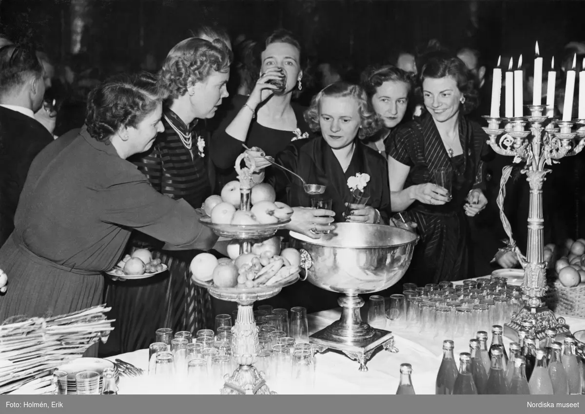 En grupp festklädda kvinnor tar för sig ur en bålskål vid ett elegant dukat bord. Man firar Nordiska kompaniets 50-årsjubileum i januari 1952. Lokalen är sannolikt Stockholms stadshus.
