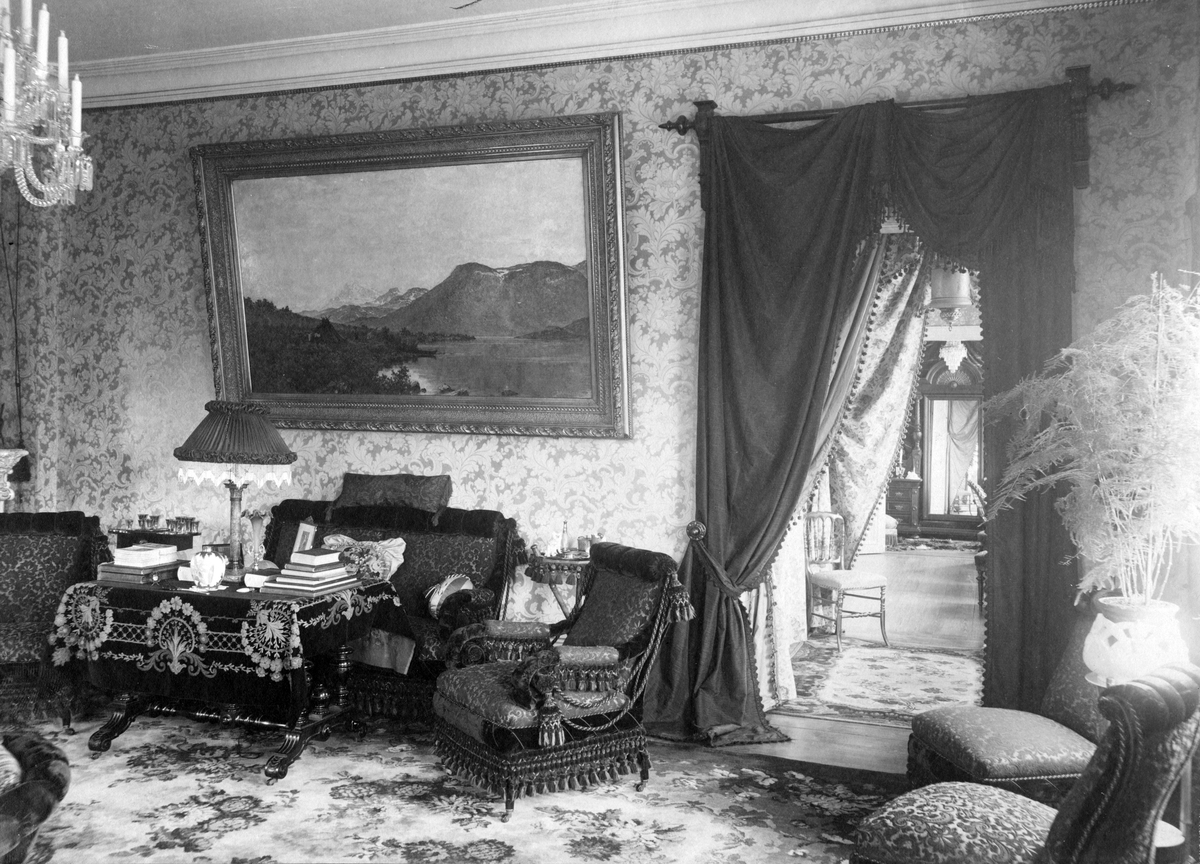Interiörbild av herrgården i Kolsva. Stoppade möbler, draperier och en stor tavla med fjällmotiv pryder rummet. Omkring 1900.