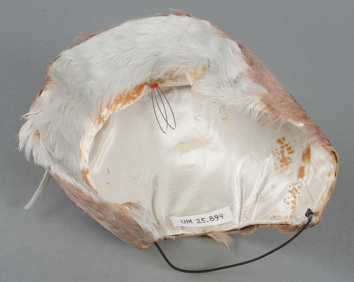 Hatt tillverkad av lilafärgade dun klädd på stramaljliknande material. Invändigt sidentyg. Liten stålfjäder fäster huvudprydnaden vid huvudet. Gummisnodd under hakan.
