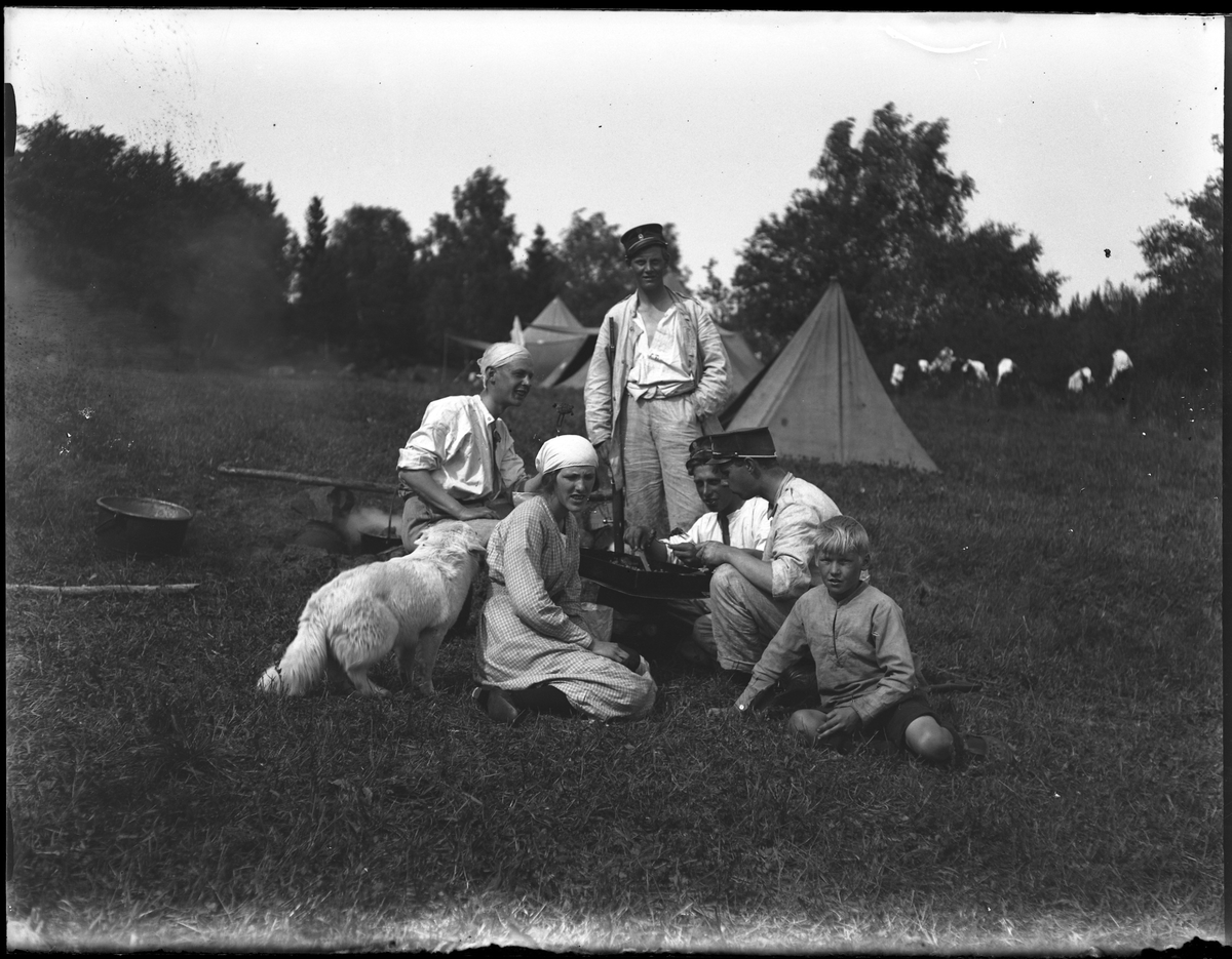 Gruppbild tagen under fotografen Harald Olssons excercistid. Framför några tält sitter en kvinna, en pojke, en hund och tre män runt ett friluftskök. Bakom dem står en man i uppknäppt sommaruniform och med ett vapen i handen.