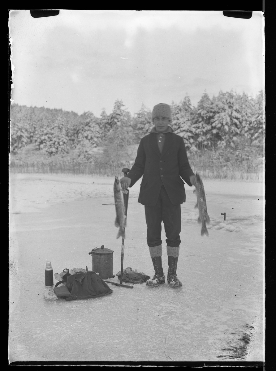 En man står på isen med två fiskar i handen, bredvid honom ligger redskap och fika. I fotografens egna anteckningar står det "Angelfiske på Kvarnsjön"