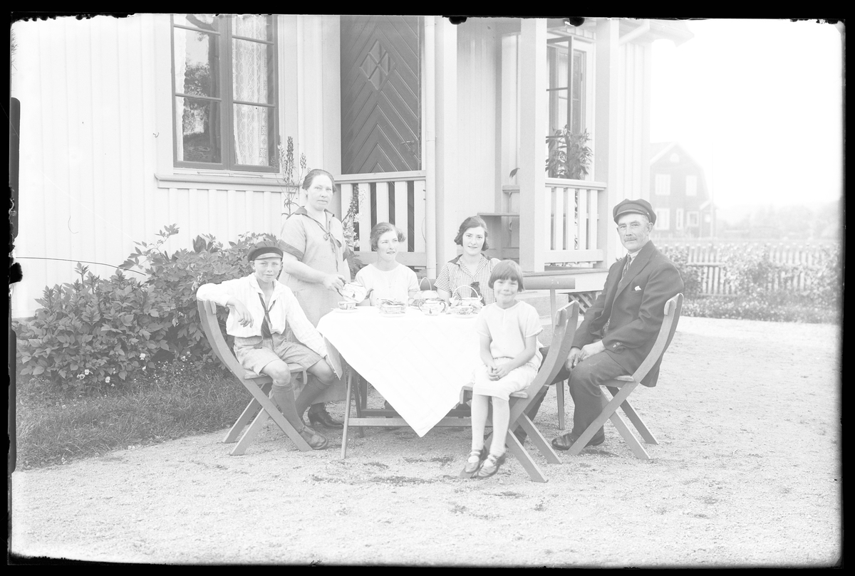 I en trädgård sitter fyra barn och en man vid ett dukat fikabord medan en kvinna står bredvid och serverar. I fotografens anteckningar står "Familjen Karl Johansson".